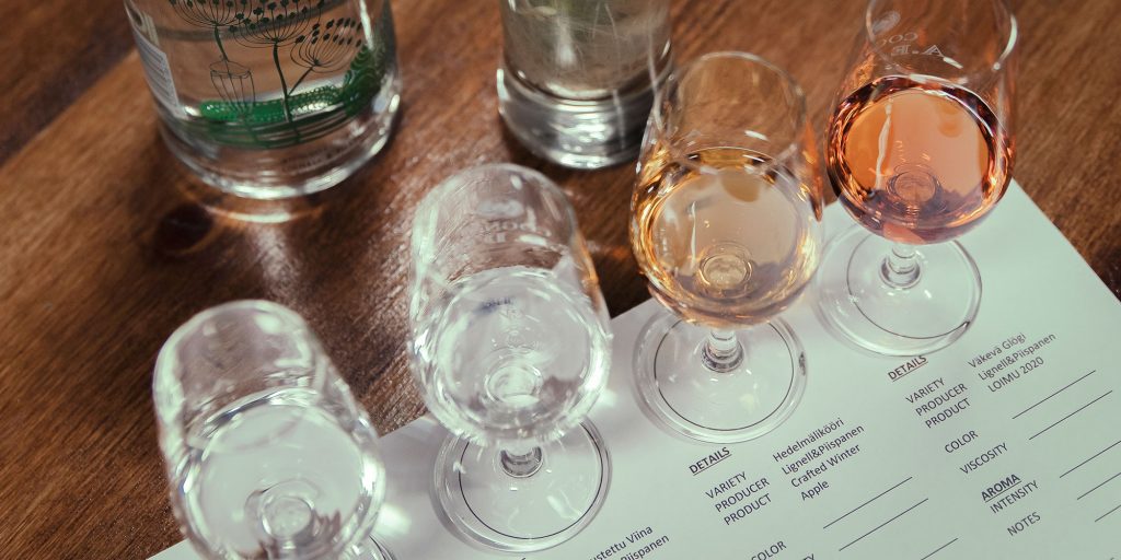 Kuvassa rivissä neljä lasia, joissa on erilaisia alkoholijuomia. Lasien alla on paperi, johon voi tehdä muistiinpanoja.