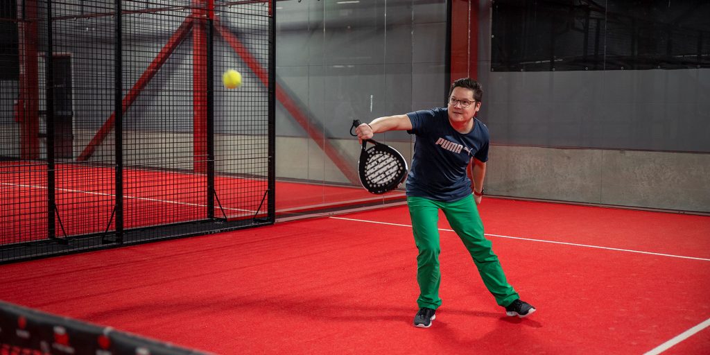 Kuvassa vihreähousuinen ihminen lyö mailalla palloa. Taustalla betoniseinää ja punainen padelkentän lattia.