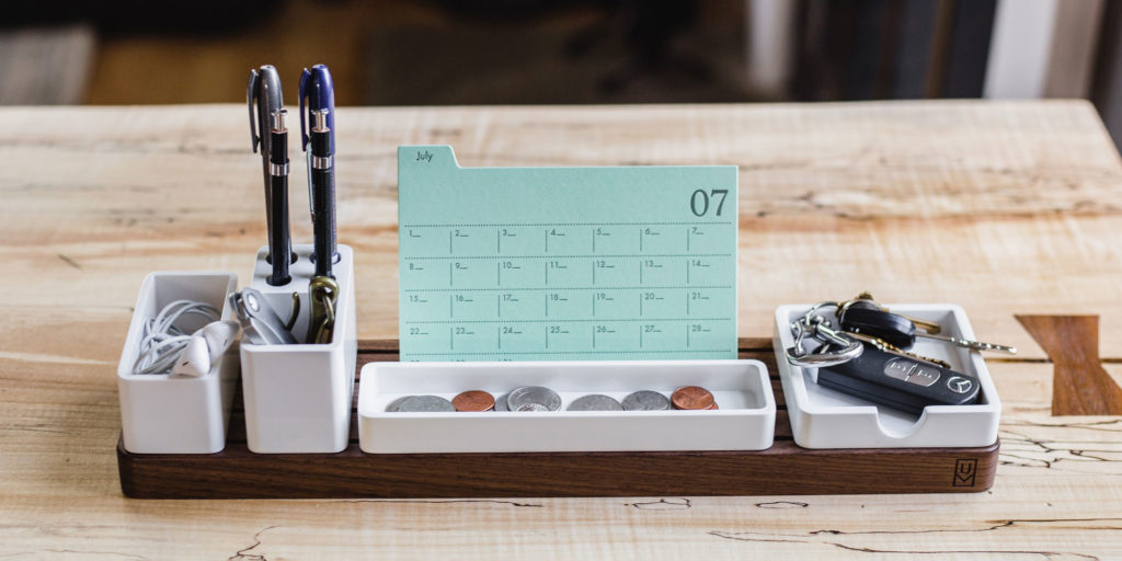 Kuvassa on puisella pöydällä kynäteline jossa on kaksi kynää, kalenteri, kolikoita, avaimet ja nappikuulokkeet.