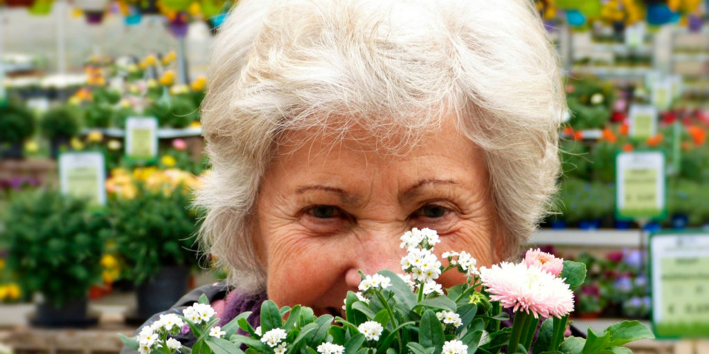 Kuvassa valkohiuksinen nainen katsoo kameraan kukkakimpun takaa.